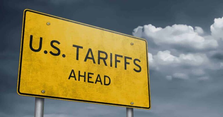 A yellow road sign with black text reads "U.S. TARIFFS AHEAD" against a cloudy, dramatic sky.