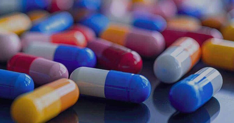 A dark, high-resolution macro shot showing a pile of multicolored pharmaceutical capsules (red, white, blue, orange, and pink), representing the branded drug market and the fragility of the supply chain.
